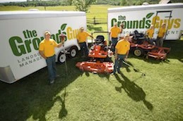 The Grounds Guys employees posing with lawn equipment in front of two company trailers.