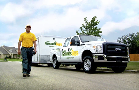 Grounds Guys service professional walking next to branded company truck and trailer.