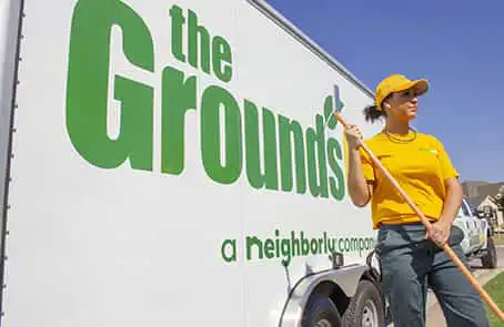 Grounds Guys employee wearing yellow hat and shirt, holding rake and standing next to branded company trailer.