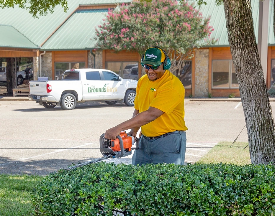 Grounds Guys employee trimming hedges, with branded company truck parked in lot behind him.