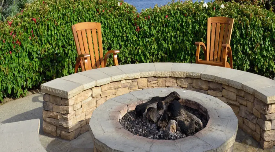 Stone firepit with semicircle stone bench, two Adirondack chairs, and hedges in the background.