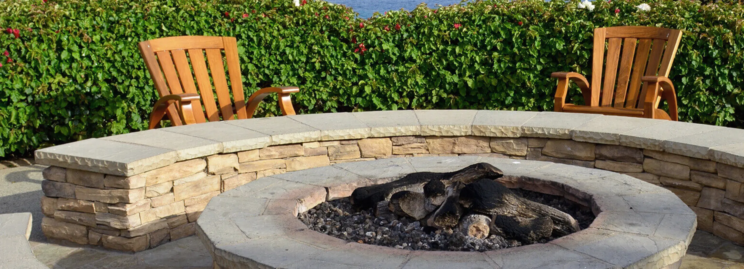 Stone firepit surrounded by low wall with pair of wooden patio chairs and hedge in background.