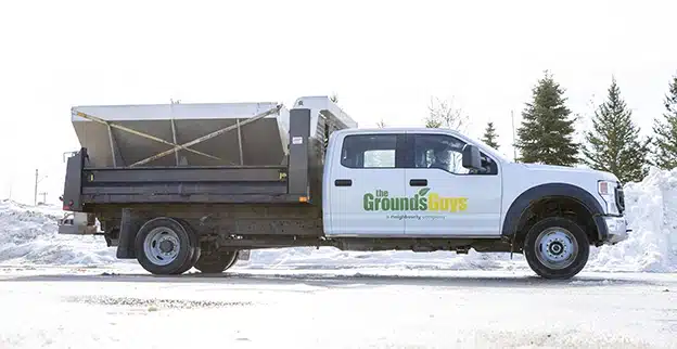 A salt truck, branded with The Grounds Guys logo, in a parking lot cleared of snow.