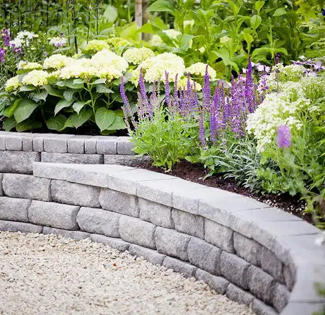 A raised plant bed with lush flowers and a stone-block retaining wall, framing a gravel path.