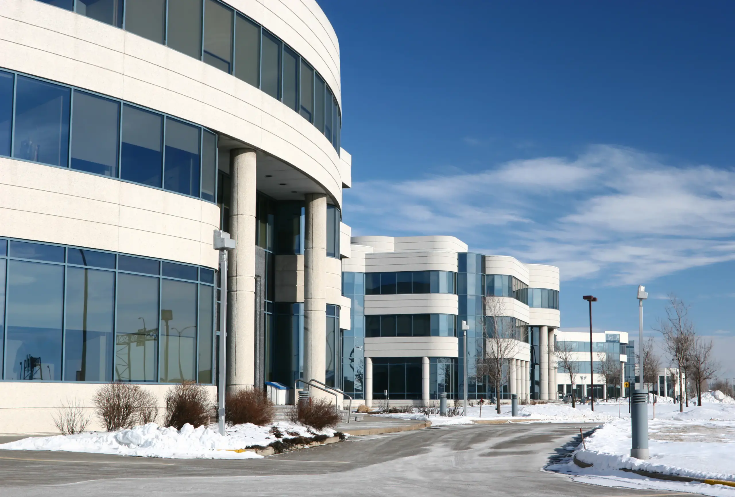 A commercial office park with multiple glass-windowed buildings in winter, with snow covering its grounds while its sidewalks have been cleared for foot traffic.
